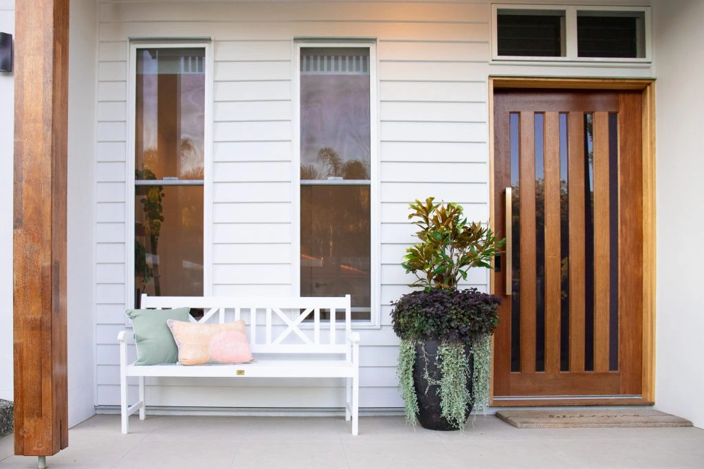 Close up photo of entry to home with large timber door, big potted plant and outdoor lounge with cushions