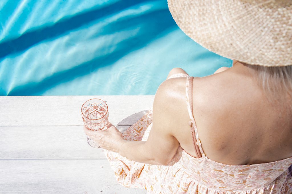 A close up view of a woman sitting beside a pool in a large hat with a pink glass