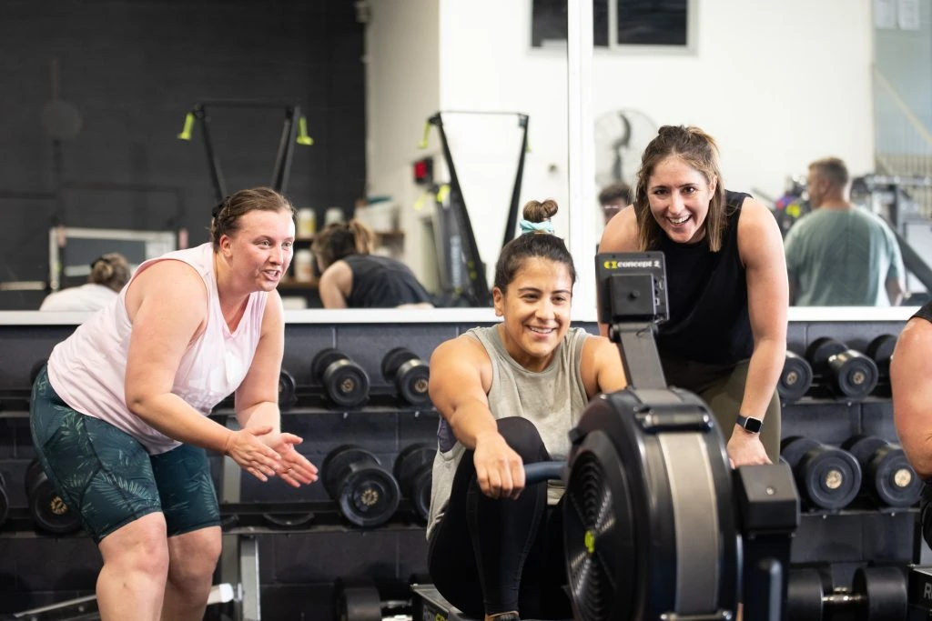 Three women at a gym, one is on a rower while being cheered on by the other two women.