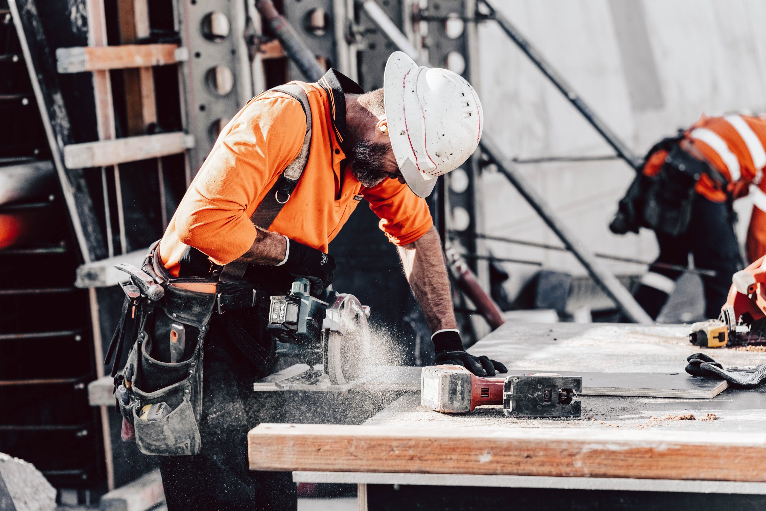 A photograph of a man in high vis and a hard hat leaning against a bench using a drop saw on a building site. The saw is kicking up dust.