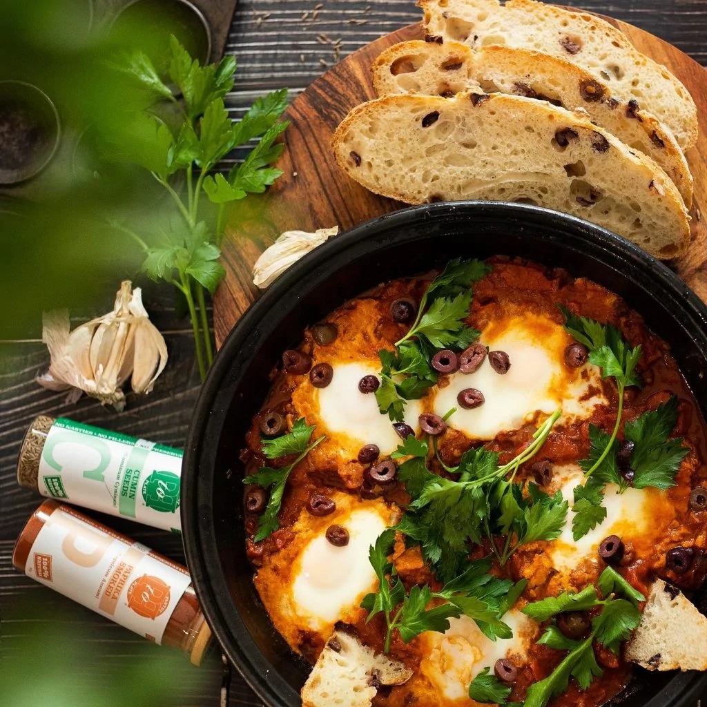 A large plate of shakshuka with two spice jars and olive sour dough.