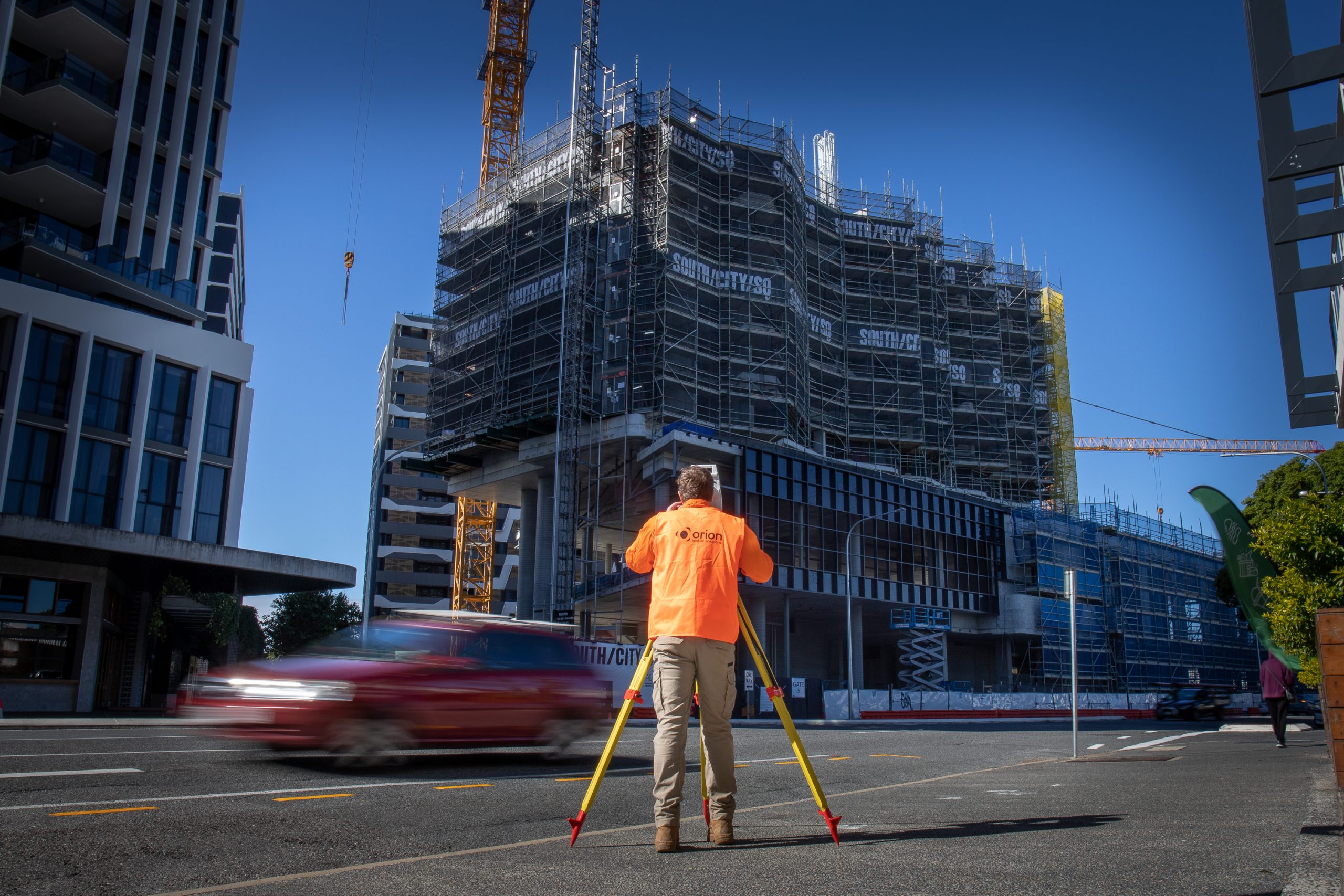 A surveyor in high vis orange looking towards a building under construction with a blurred car driving past.
